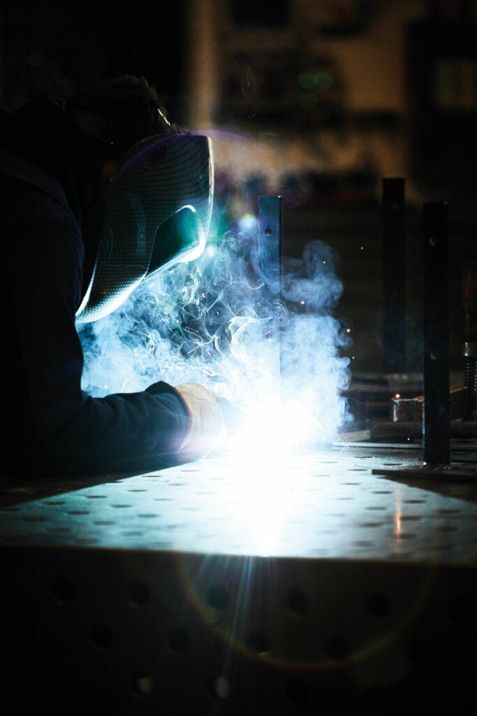 Schweißer bei der Arbeit an metallener Werkbank, umgeben von Funken.