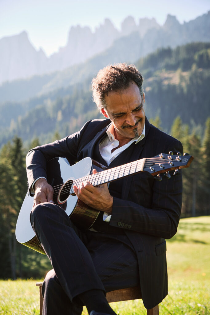 Mann in Anzug spielt Gitarre vor Bergkulisse im Freien