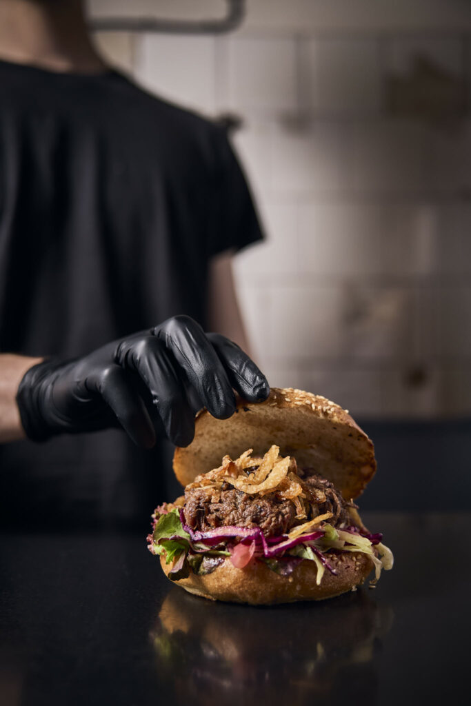 Person mit Handschuh richtet Burger mit Gemüse und Fleisch an.