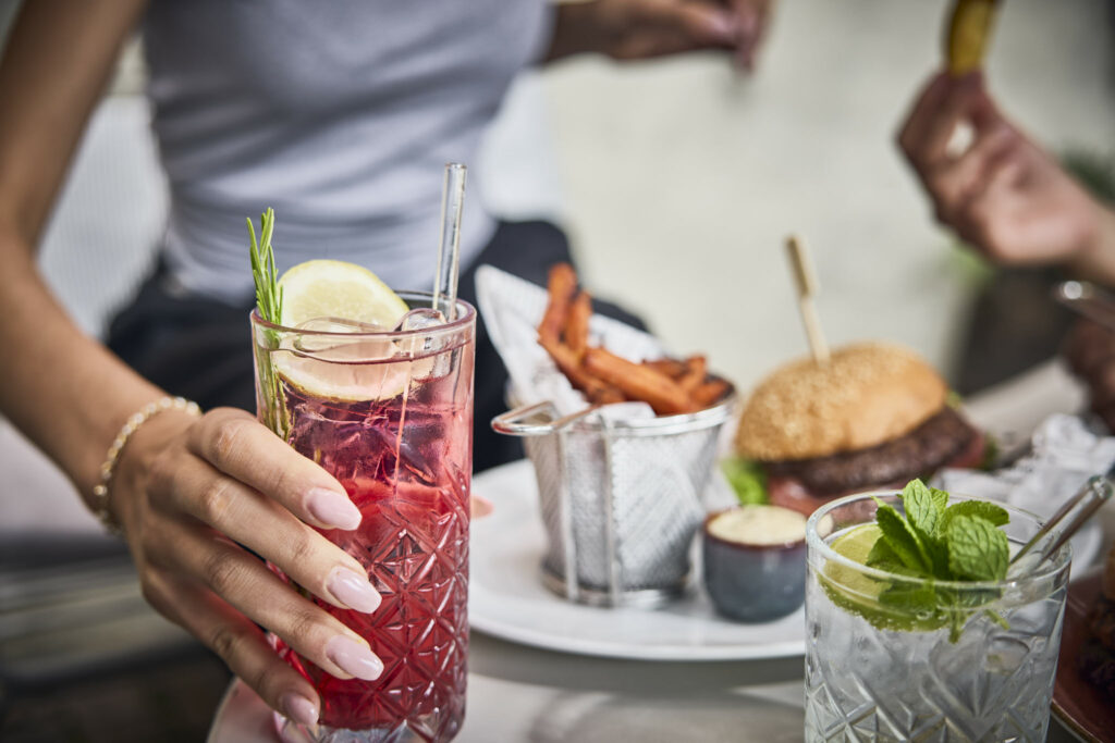 Frau hält roten Cocktail, Burger und Pommes im Hintergrund.