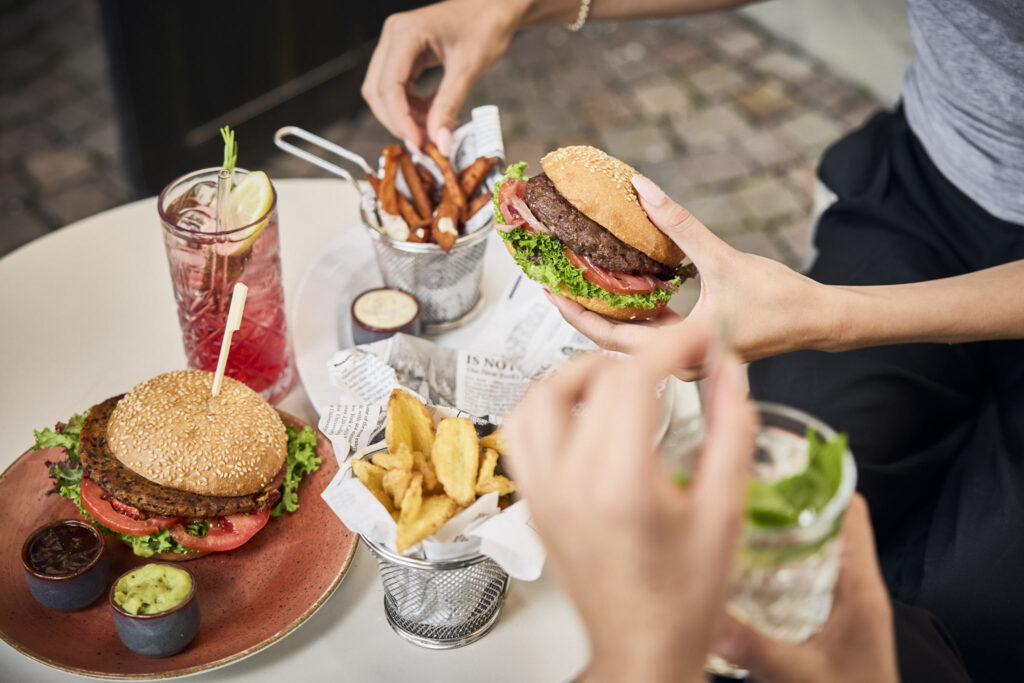 Personen essen Burger und Pommes an kleinem Tisch im Freien.