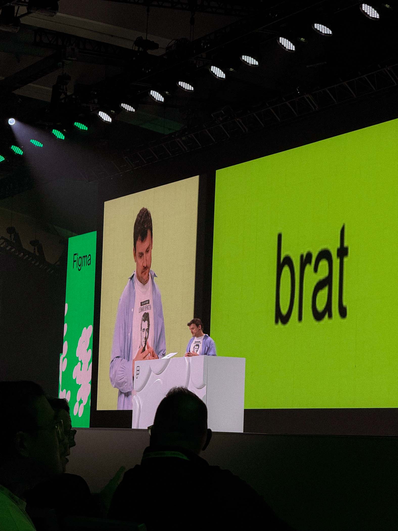 Mann hält Präsentation auf großer Bühne, grüner Bildschirm mit 'brat